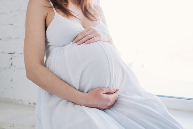 A woman in a white dress hugging her pregnant stomach against a white background
