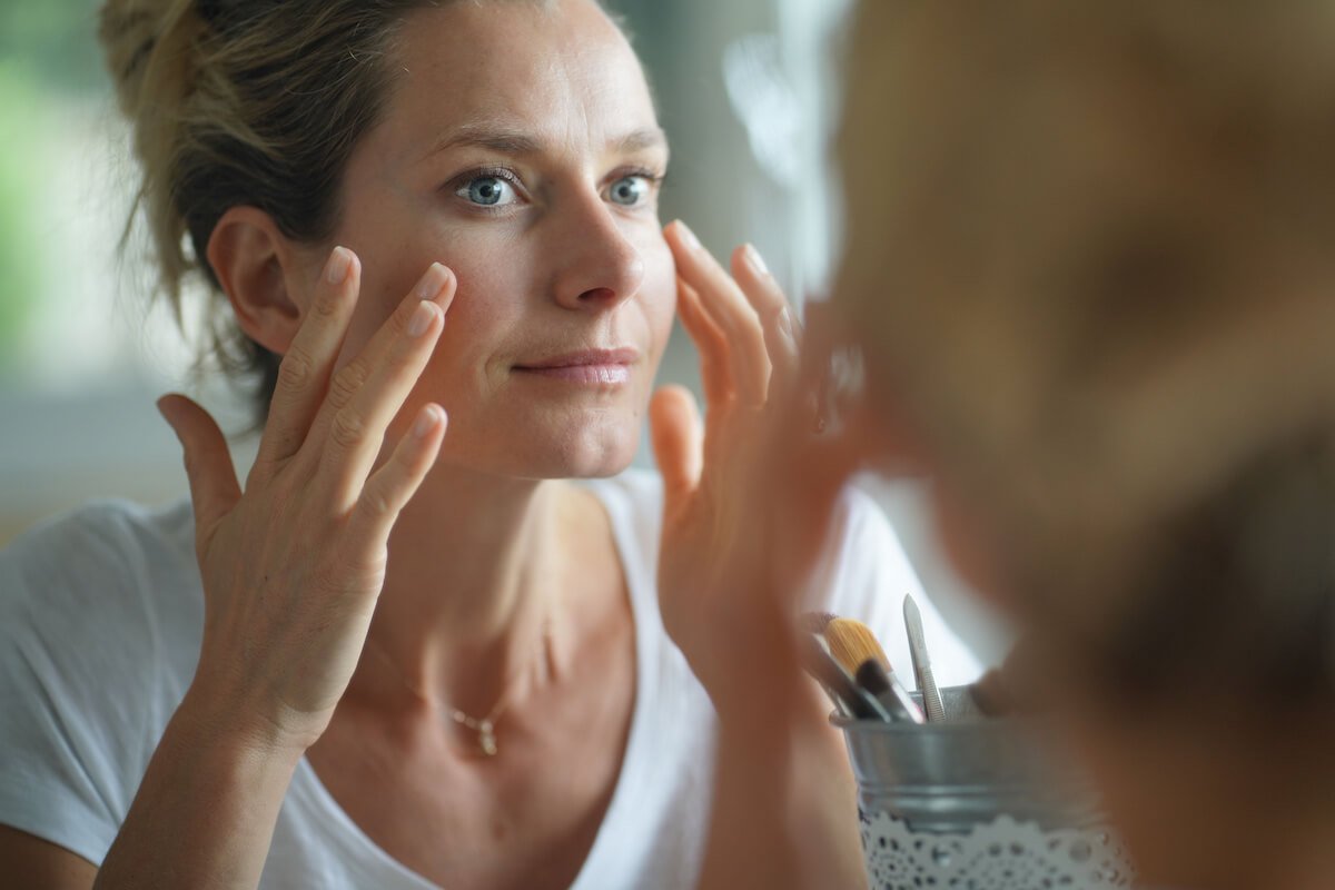 Woman with hair pulled back touching her face in the mirror