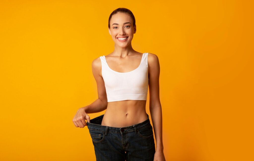a lady on a light orange background holding her loose pants while smiling