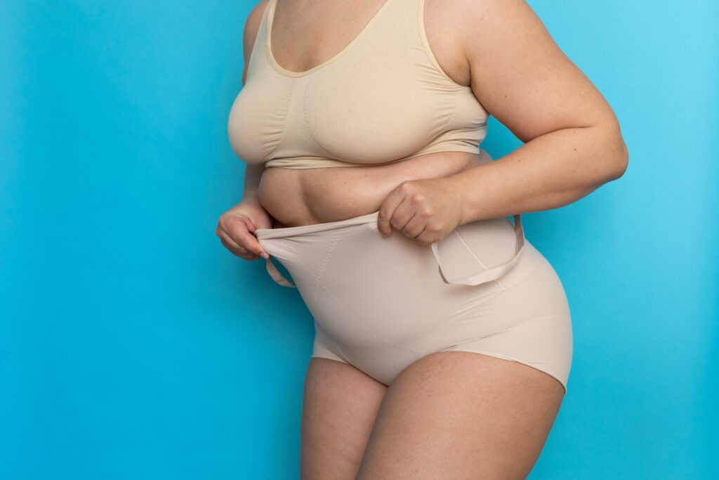 A overweight women with light blue background holding her undergarment over her waist