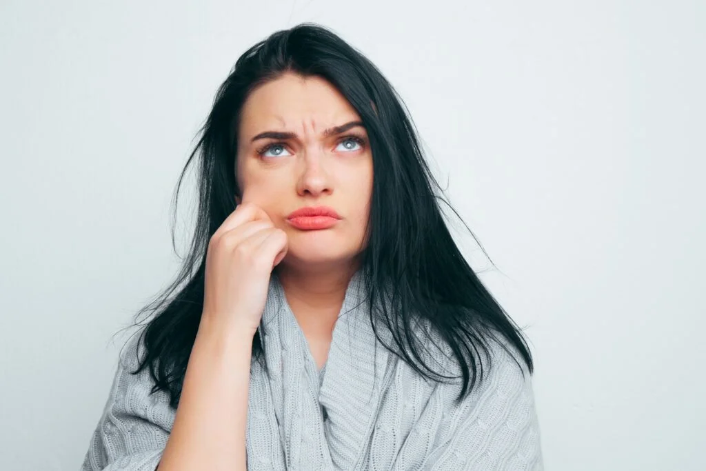 A young beautiful girl in a gray robe pinching her cheek, thinking about fat cheeks, about excess weight, and that it is time to lose weight on a gray-white studio background.