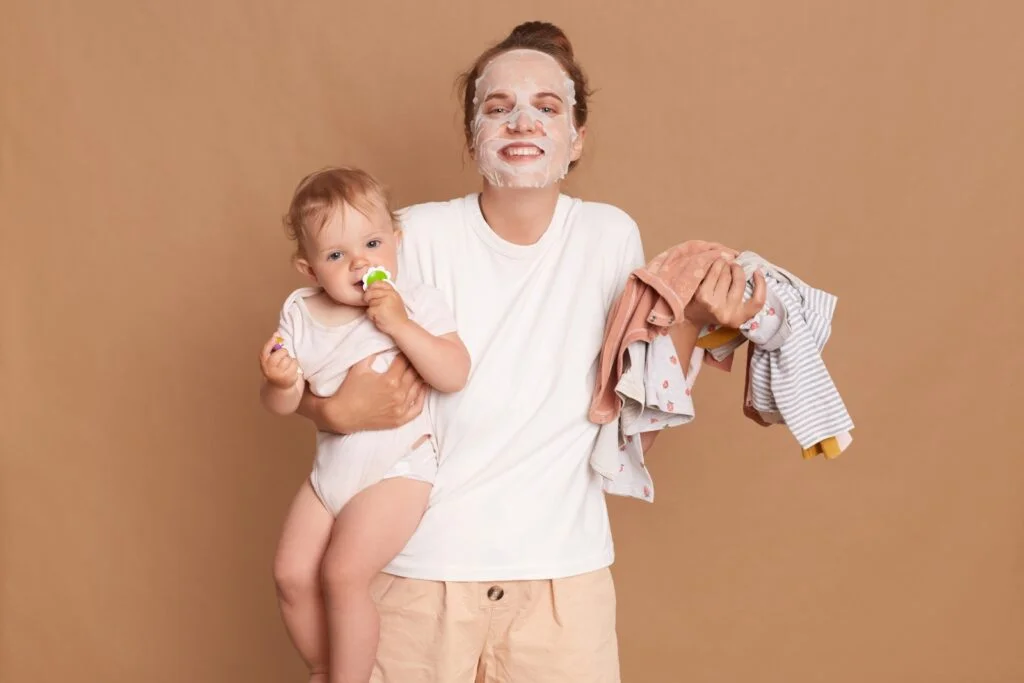 Happy positive woman wearing white t shirt doing cosmetic procedures while playing with kid and doing household chores, holding baby daughter in hands, expressing positive emotions.